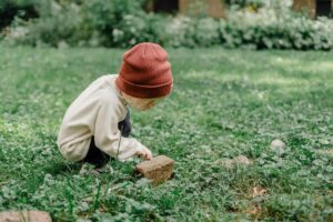 Full body side view of playful kid wearing hat squatting on grassy lawn while exploring small stone against blurred background