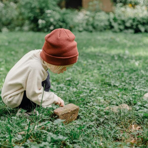 Full body side view of playful kid wearing hat squatting on grassy lawn while exploring small stone against blurred background