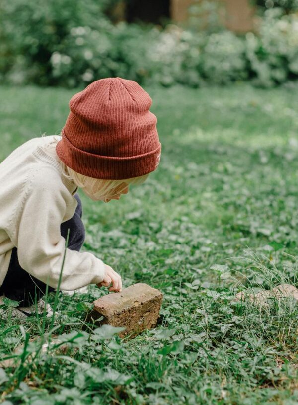 Full body side view of playful kid wearing hat squatting on grassy lawn while exploring small stone against blurred background